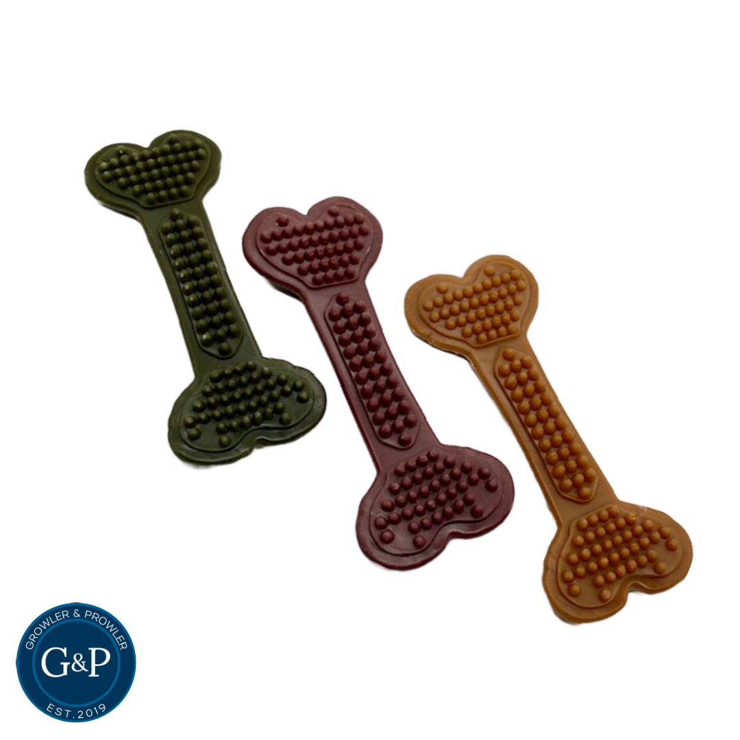 Three bone-shaped dog chews in green, brown, and purple. The chews have raised nubs for dental cleaning and the Growler & Prowler logo and 'EST. 2019' printed on them